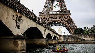 A rescue boat cruises on the Seine river beneath the Eiffel Tower, adorned with the Olympic rings.