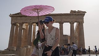 Touristen schützen sich vor der Hitze mit einem Spaziergang unter einem Regenschirm am Parthenon auf der antiken Akropolis im Zentrum Athens im Juni - dem heißesten Juni aller Zeiten