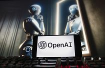 The OpenAI logo is displayed on a cell phone with an image on a computer monitor