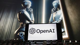 The OpenAI logo is displayed on a cell phone with an image on a computer monitor