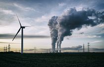 Un aerogenerador, una central eléctrica de carbón con contaminación y torres de alta tensión al fondo.