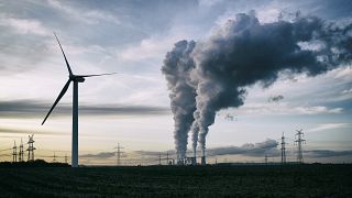 Une seule éolienne, et en arrière-plan, une centrale électrique au charbon qui pollue et des pylônes électriques.
