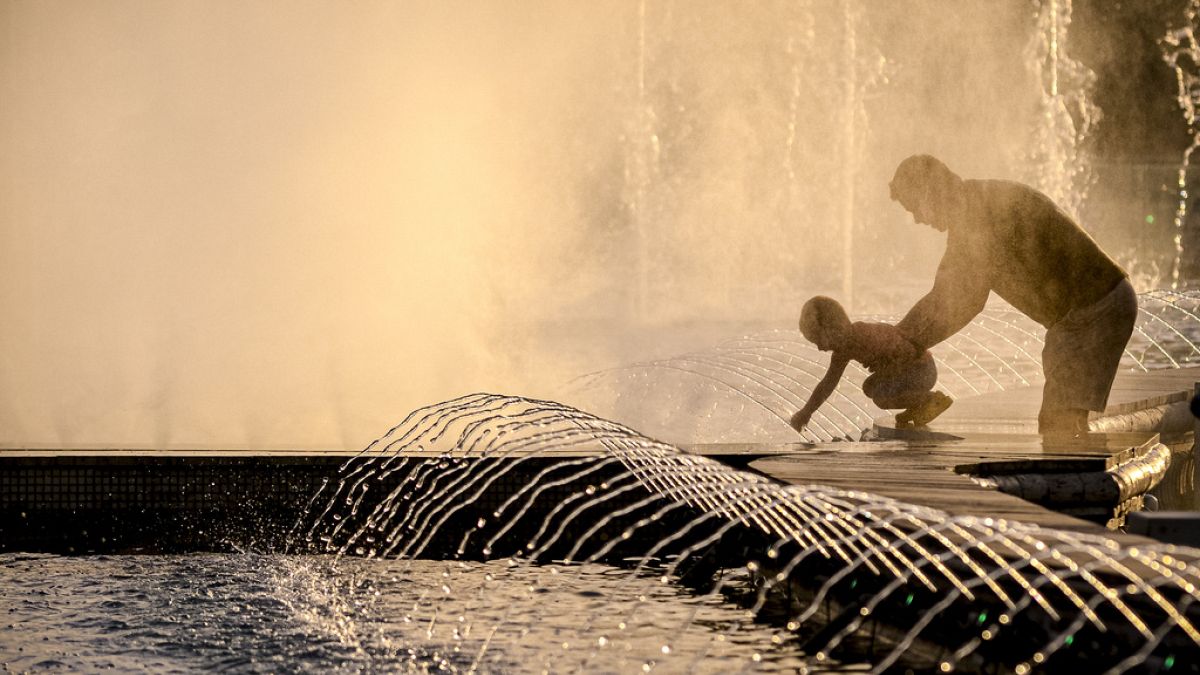 Um homem segura um bebé junto às fontes da cidade numa noite quente em Bucareste, Roménia, sexta-feira, 21 de junho de 2024, quando as temperaturas ultrapassaram os 35 graus c