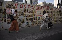 İsrail'in Tel Aviv kentinde Rehineler Meydanı olarak bilinen meydanda insanlar Gazze Şeridi'nde tutulan rehinelerin fotoğraflarının bulunduğu bir duvarın önünden geçiyor