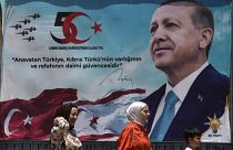 Mulheres turcas e uma criança caminham em frente a um cartaz do Presidente Recep Tayyip Erdoğan após uma parada militar no norte de Chipre, 20 de julho de 2024
