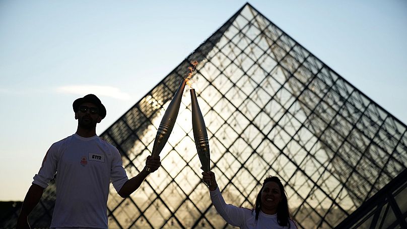 French photographer JR, left, and athlete Sandra Laoura hold the Olympic torch in the courtyard of the Louvre museum, 14 July 2024 in Paris.