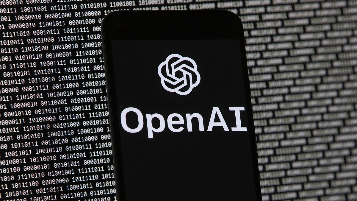 El logotipo de OpenAI aparece en un teléfono móvil frente a una pantalla de ordenador con datos binarios aleatorios.