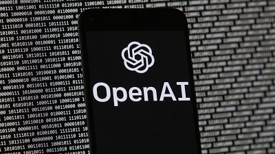 The OpenAI logo appears on a mobile phone in front of a computer screen with random binary data.