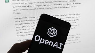 The OpenAI logo is seen on a mobile phone in front of a computer screen displaying output from ChatGPT, on March 21, 2023, in Boston