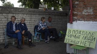 Residentes locais sentam-se perto de um abrigo em Sudzha com um cartaz que diz "Civis na cave. Sem militares", 16 de agosto de 2024