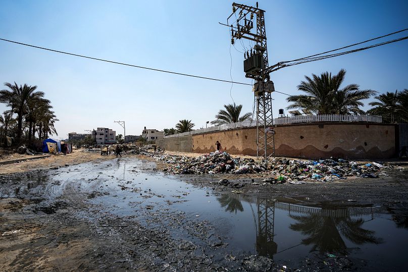 فلسطينيون نازحون بسبب الهجوم الجوي والبري الإسرائيلي على قطاع غزة يسيرون بجوار خط داكن من مياه الصرف الصحي المتدفقة في شوارع دير البلح،  الثلاثاء، 27 أغسطس/آب 2024