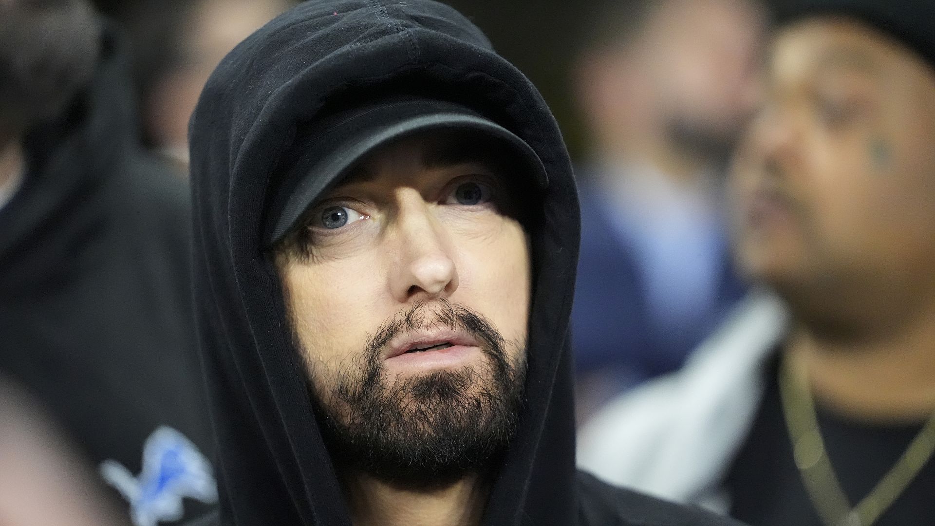 El rapero Eminem aparece en primer plano, vestido con una sudadera con capucha negra y una gorra del mismo color. Su rostro está serio, con una expresión pensativa, y su barba es visible. El fondo está desenfocado, con varias personas borrosas, lo que hace que Eminem sea el foco principal de la imagen.