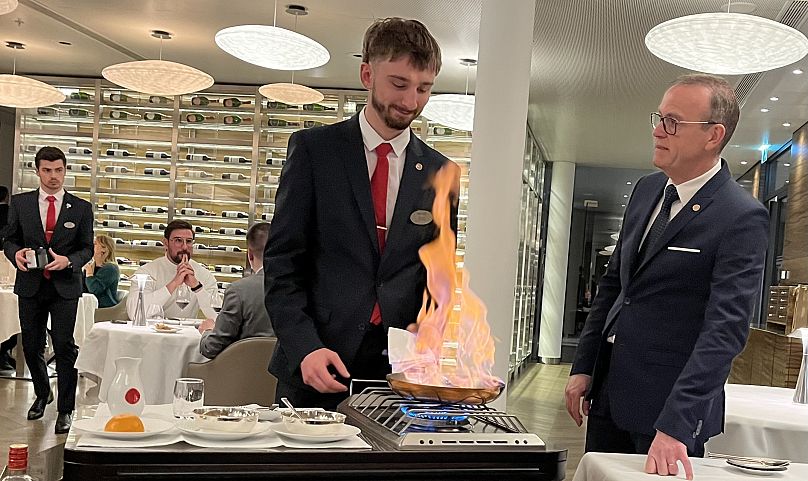 A student prepares a flambée for diners in front of supervisors.