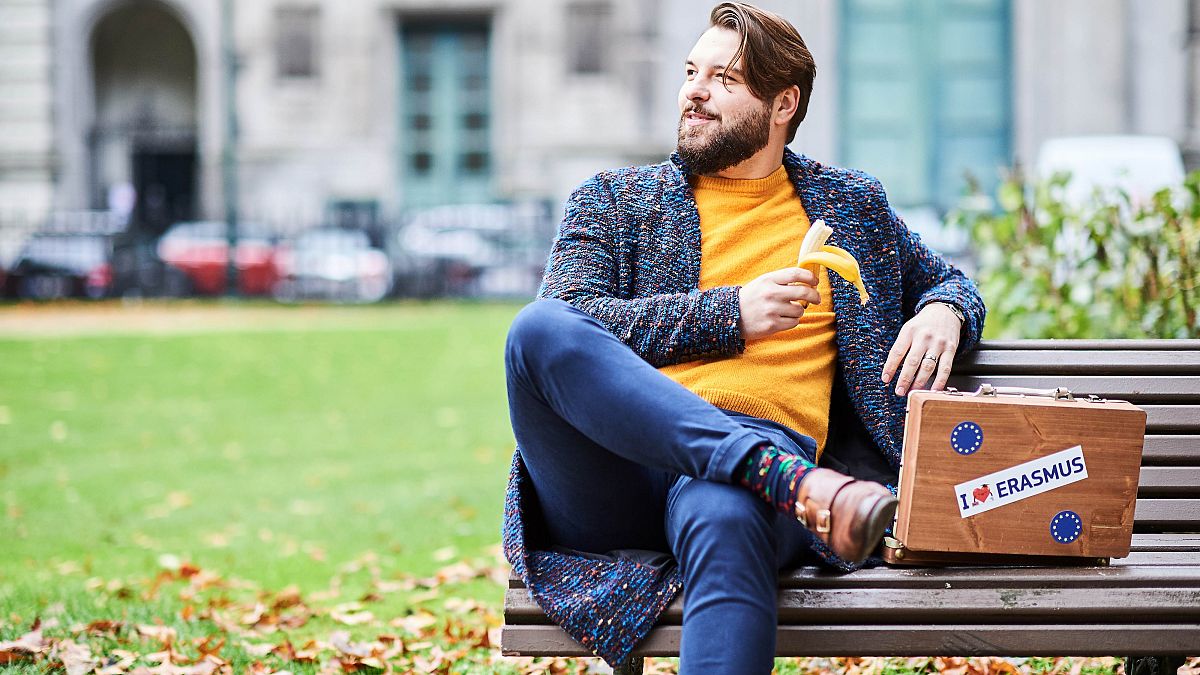 Joven en un banco con una maleta que muestra el logotipo de Erasmus (21/10/2019)