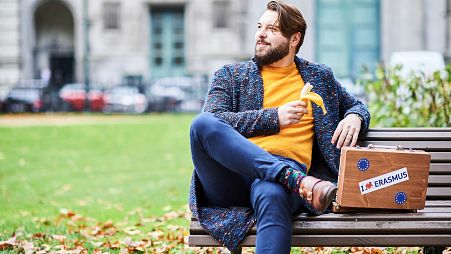 Joven en un banco con una maleta que muestra el logotipo de Erasmus (21/10/2019)