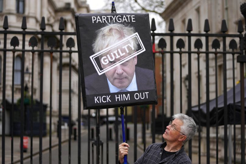 A protester holds a sign showing British Prime Minister Boris Johnson as he stands in front of the entrance to Downing Street in London, Wednesday, April 13, 2022