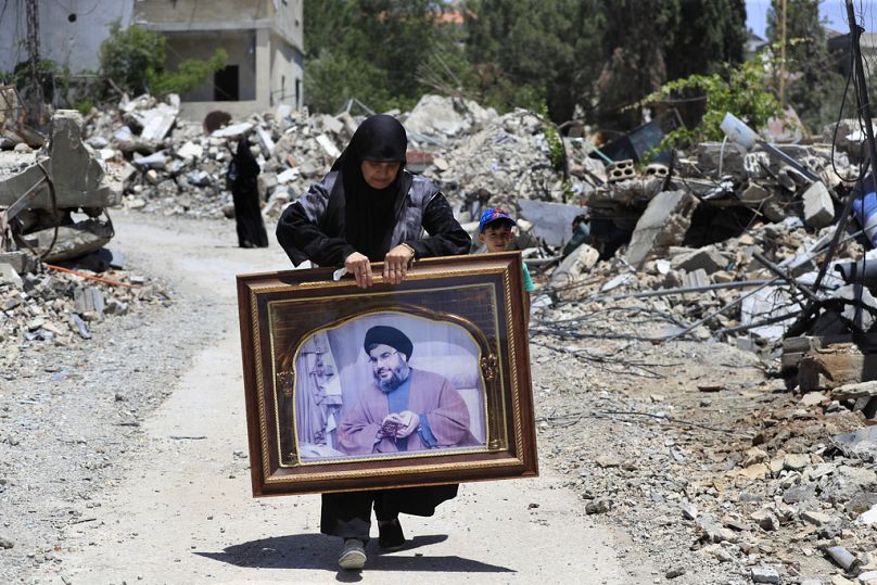 Uma mulher carrega uma moldura que mostra um retrato do líder do Hezbollah, Sayyid Hassan Nasrallah, enquanto passa por casas destruídas que foram atingidas por ataques aéreos