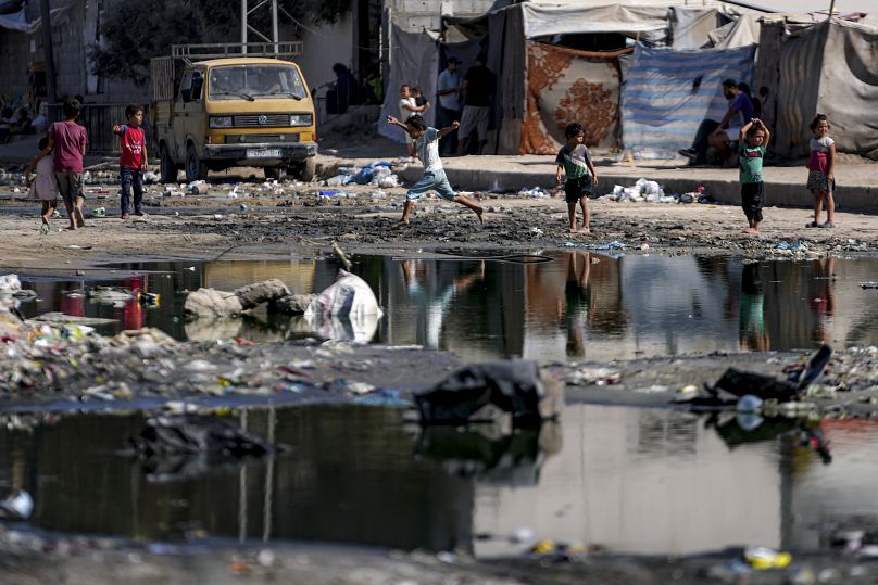 أطفال نازحون يسيرون عبر خط مظلم من مياه الصرف الصحي المتدفقة في شوارع دير البلح، وسط قطاع غزة، الخميس، 29 أغسطس/آب 2024