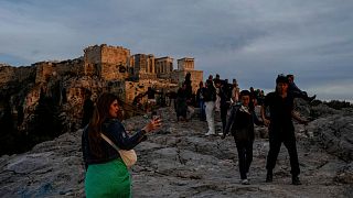 Turisti passeggiano sulla collina dell'Areopago, di fronte all'antica Acropoli, mentre il sole tramonta ad Atene.