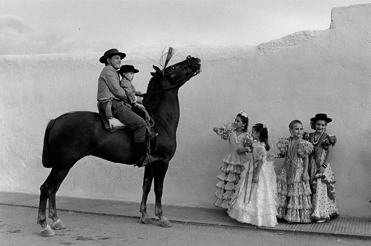 Young girls dressed in traditional Sevillana attire pose next to a horse.