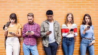 A row of young people standing against a brick wall, busy on their phones. 