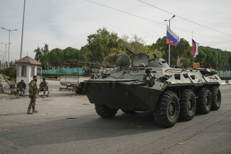 A Russian armoured vehicle drives past Syrian fighters guarding Lakatia airport in the town of Khmeimim, southeast of the city of Latakia, Syria, Monday, Dec. 16, 2024.