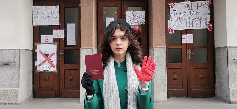 Emilija em frente à sua universidade com um livro de registo e uma luva vermelha.