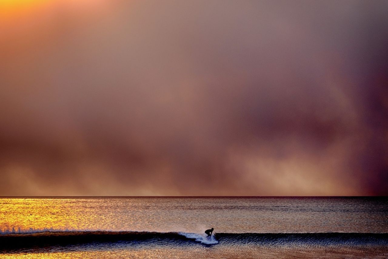 A surfer takes off on a wave in Santa Monica, Calif., during sunset under a blackened sky from the Palisades fire in the Pacific Palisades on Jan. 7, 2025.