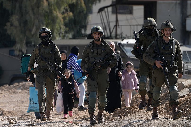 جنود إسرائيليون يسيرون أمام فلسطينيين نزحوا من مخيم جنين للاجئين في الضفة الغربية وهم يحملون أمتعتهم يوم الخميس، 23 يناير/كانون الثاني 2025