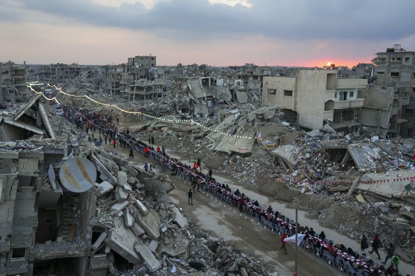 As the sun sets, Palestinians sit at a large table surrounded by the rubble of destroyed homes and buildings as they gather for iftar, the fast-breaking meal, on the first day