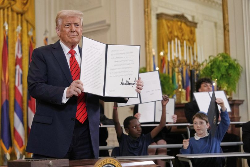 Trump holds up a signed executive order as young people hold up copies of the executive order they signed at an education event in the White House's East Room, March 20, 2025