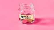 A glass jar with some coins and a label saying "Budget" against a pink background.