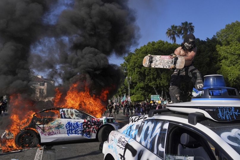 Ein Polizeiwagen wird mutwillig zerstört, während ein anderer in der Nähe des Metropolitan Detention Centers in der Innenstadt von Los Angeles brennt. Sonntag, 8. Juni 2025
