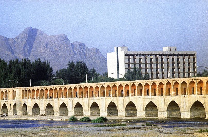 جسر الثلاثة والثلاثين قوسًا"، الذي بُني عام 1602، يتباين مع مبنى مكاتب حديث وقمم جبل كوه إي سوفه المُسننة في أصفهان، إيران، يوليو 1971. 