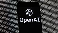 The OpenAI logo appears on a mobile phone in front of a computer screen with random binary data, March 9, 2023, in Boston