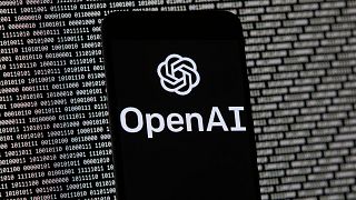 The OpenAI logo appears on a mobile phone in front of a computer screen with random binary data, March 9, 2023, in Boston