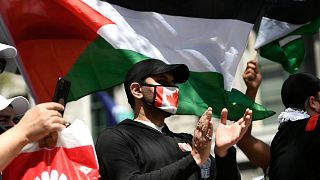 فردی با ماسک پرچم کانادا در جریان تجمع اعتراضی در اتاوا در ۱۵ مه ۲۰۲۱ علیه حملات نظامی اسرائیل به سرزمین‌های فلسطینی که در پشت سرش پرچم فلسطین دیده می‌شود