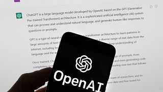 The OpenAI logo is seen on a mobile phone in front of a computer screen which displays output from ChatGPT, March 21, 2023