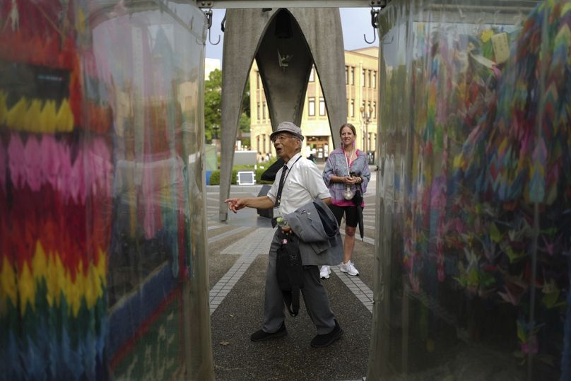 Kunihiko Iida, an atomic bomb survivor and volunteer guide, speaks English to foreign visitors in front of the Children's Peace Monument, where people bring paper cranes.