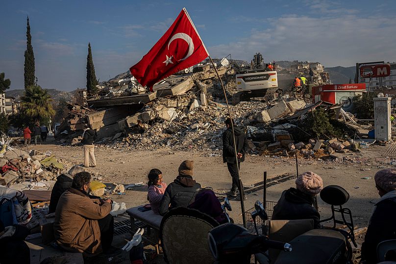 A family sit next to a collapsed building as they wait for the bodies of their relatives to be recovered from under the rubble in Antakya, southeastern Turkey, February 2023.