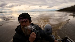 Jake Pogrebinsky fuma un cigarrillo mientras navega con su barca en el río Yukón en busca de troncos para recolectar, el martes 20 de mayo de 2025, en Galena, Alaska.