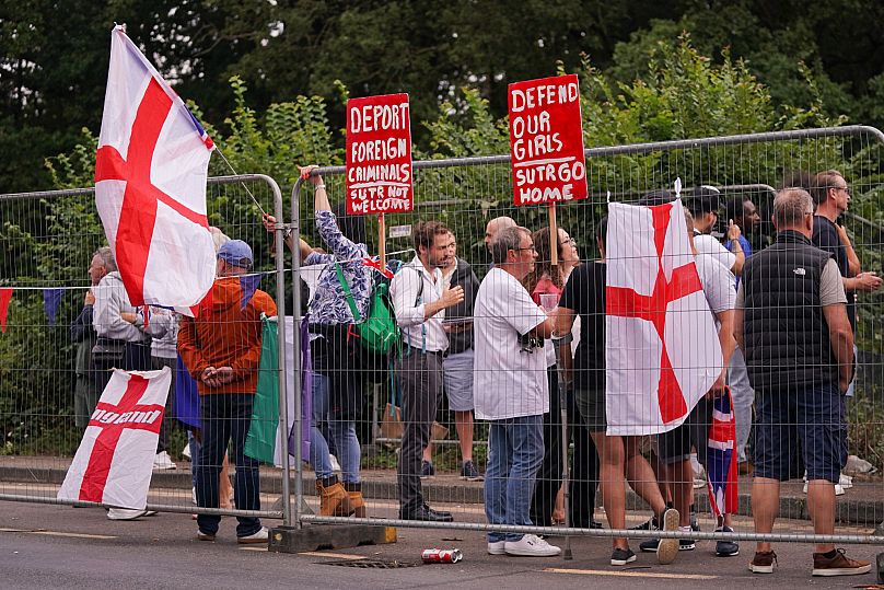 Demonstrators wave England and Union flags as they stand outside the Bell Hotel in Epping, 27 July, 2025
