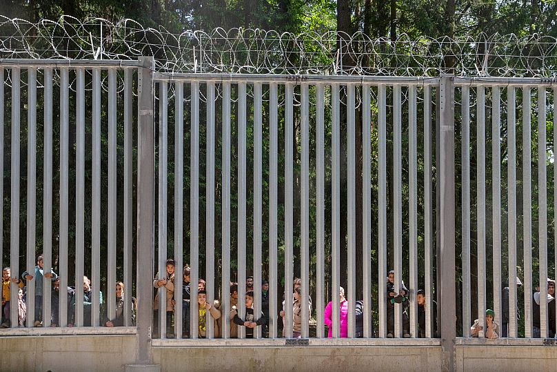 A group of some 30 migrants seeking asylum look through the railings of a wall that Poland has built on its border with Belarus in Bialowieza, 28 May, 2023