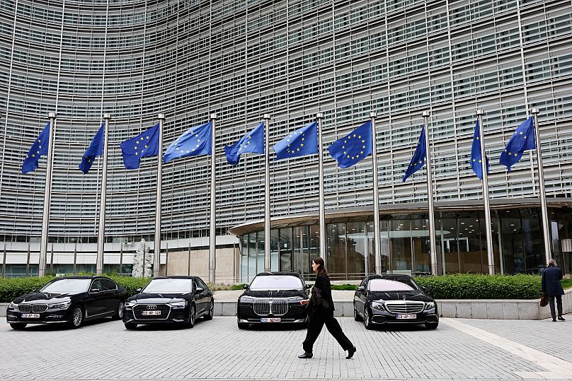 Οι σημαίες της Ευρωπαϊκής Ένωσης ανεμίζουν στον άνεμο καθώς μια γυναίκα περνάει μπροστά από διπλωματικά οχήματα έξω από την έδρα της ΕΕ στις Βρυξέλλες, 27 Μαΐου 2025